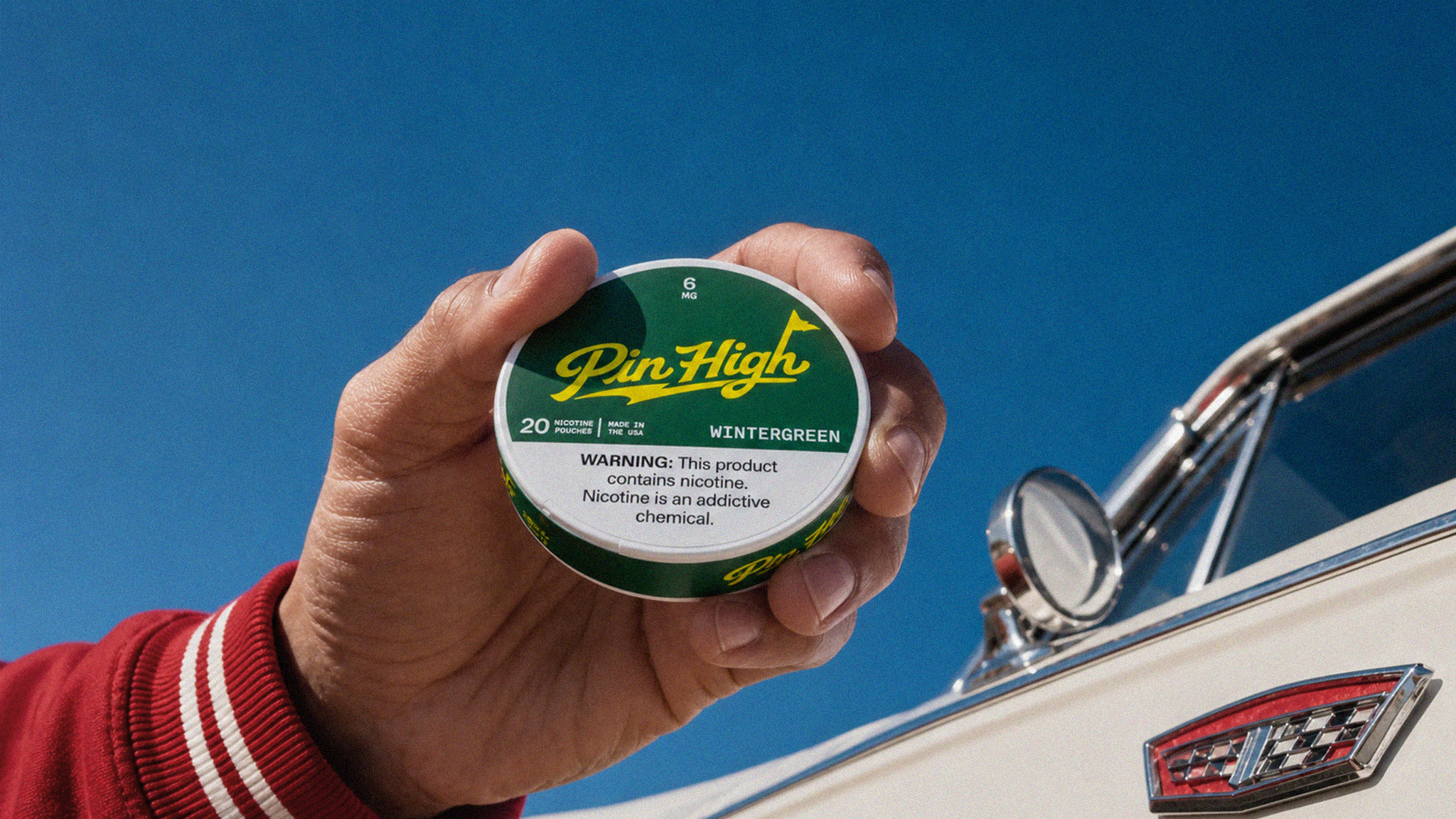 Hand holding a container of Pin High Wintergreen against a clear blue sky with a vintage car in the background.